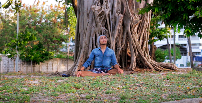 Pessoa sentada com as pernas cruzadas em borboleta, na frente de uma grande árvore (apenas o tronco presente na imagem)