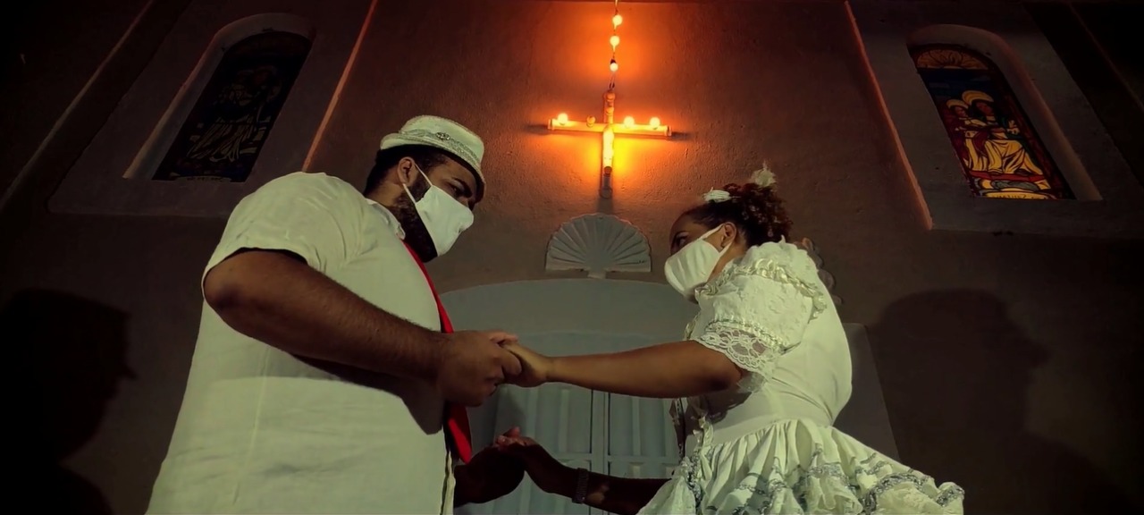 casal de noivos (homem e mulher) vestidos com roupas juninas na cor branca, usando máscaras, segurando as mãos um de frente para o outro.