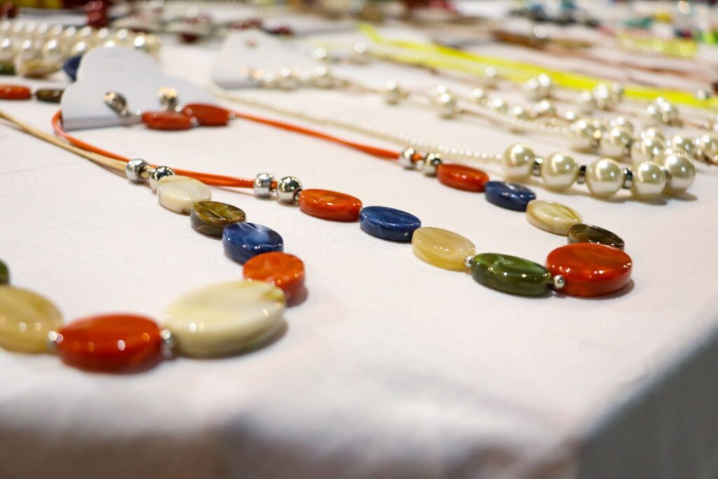 A imagem mostra uma mesa coberta por um tecido claro onde estão expostos diversos colares artesanais. Em primeiro plano, há colares feitos com contas ovais e lisas em tons vibrantes como laranja, creme, azul, verde e marrom, intercaladas por pequenas esferas metálicas. Ao fundo, um pouco desfocado, é possível ver outras peças de bijuterias, incluindo colares de pérolas e acessórios coloridos, sugerindo uma feira ou exposição de artesanato. O conjunto transmite sensação de cuidado manual e variedade de estilos.