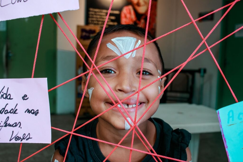Criança sorrindo em primeiro plano, com pintura branca no rosto, vista por entre uma teia de fios vermelhos esticados. Ao redor, bilhetes coloridos escritos à mão estão presos aos fios. O ambiente ao fundo parece ser uma sala com paredes verdes e móveis simples, levemente desfocado.